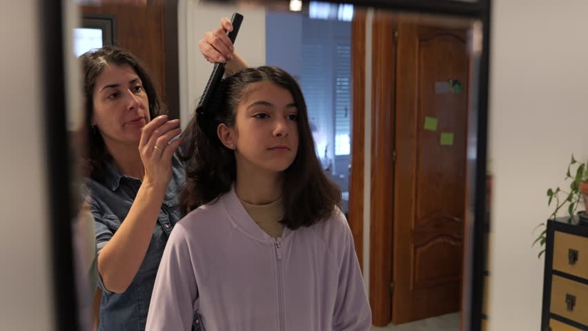 Mother combs her daughter’s hair in front of the mirror in their home in a daily routine scene preparing for school