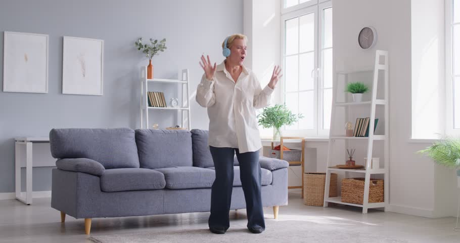Happy carefree middle aged woman dancing at home, 50s elderly business lady having fun alone listening to good music, energetic moving in modern living room interior, enjoy freedom, active lifestyle