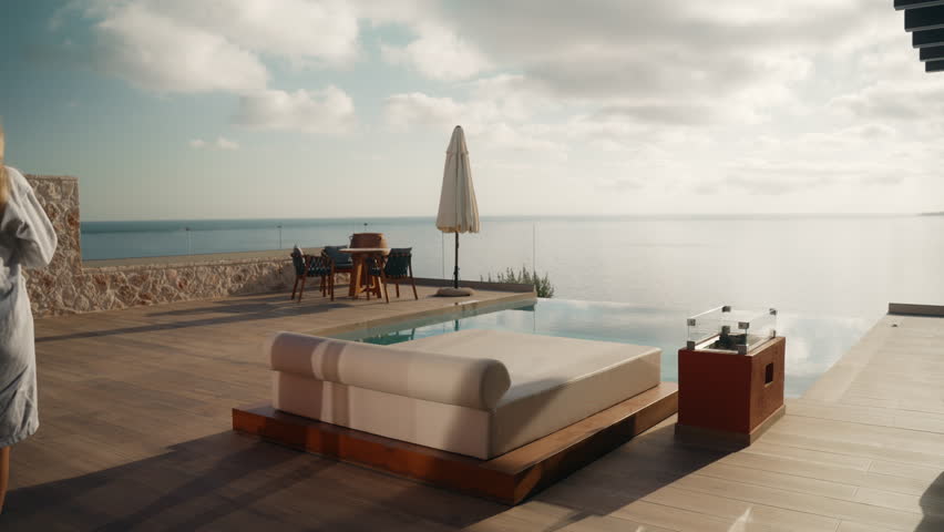 Young Blonde Woman in White Bathrobe Walking to Sunbed to Read a Book with Infinity Pool and Sea View in the background at a Luxury Villa in Greece