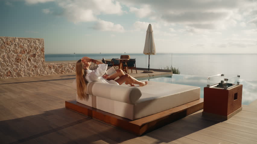 Woman in White Bathrobe Sitting on Sunbed Reading a Book with Infinity Pool and Sea View in the background