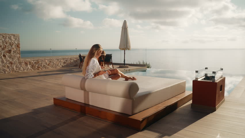 Woman in White Bathrobe Sitting on Sunbed Reading a Book with Infinity Pool and Sea View in the background