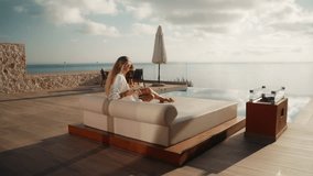 Woman in White Bathrobe Sitting on Sunbed Reading a Book with Infinity Pool and Sea View in the background - Powered by Shutterstock - Get 15% off with code: PIKWIZARD15