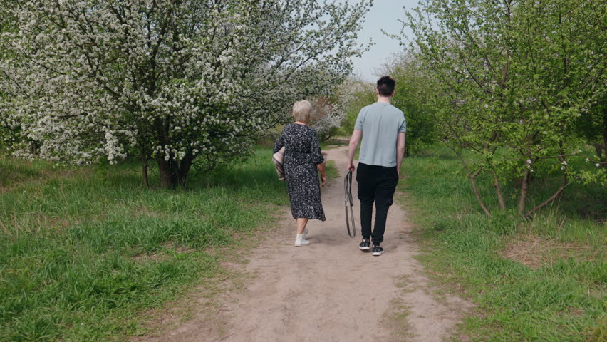 Elderly woman and her grandson walking together along a blooming garden path on a sunny spring day, with a golden retriever running nearby and a picnic blanket in hand. High quality 4k footage