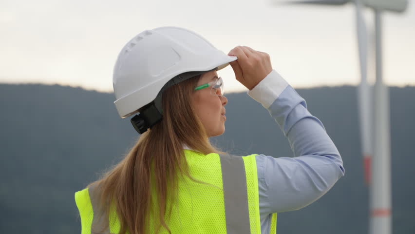 A female engineer in a hard hat and reflective vest inspects a large wind turbine, showcasing her commitment to renewable energy and sustainable development in a beautiful natural landscape