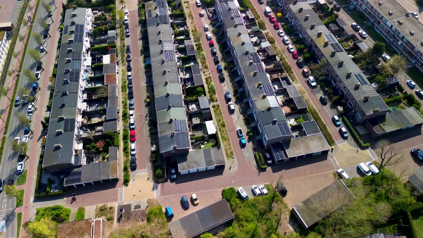 Aerial view of a residential neighborhood with rows of houses, solar panels on rooftops, parked cars along narrow streets, and small gardens, captured on a sunny day.