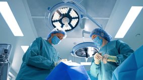 Two female medics stand at the operating table. Surgeon uses a tool performing surgery. Scrub nurse holds an instrument. Low angle view. - Powered by Shutterstock - Get 15% off with code: PIKWIZARD15