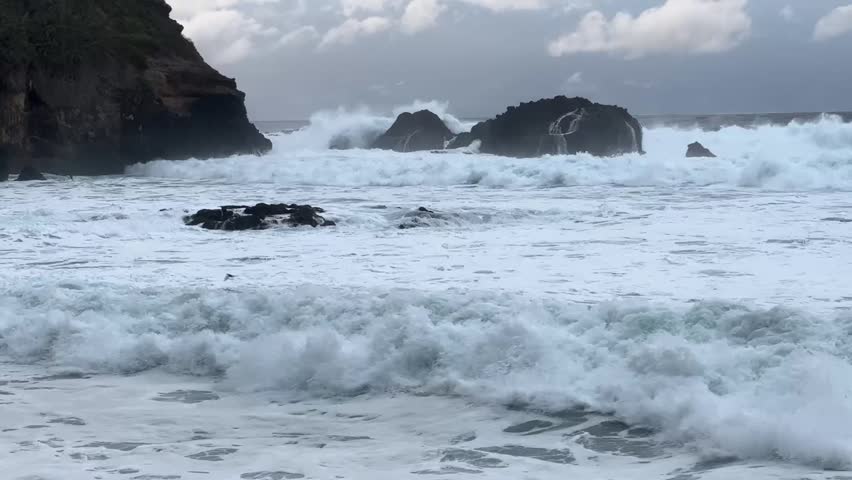Dramatic ocean waves crashing against dark volcanic rock formations during stormy weather. Powerful surf with white foamy water strikes coastal cliffs  under cloudy sky.  - Powered by Shutterstock - Get 15% off with code: PIKWIZARD15