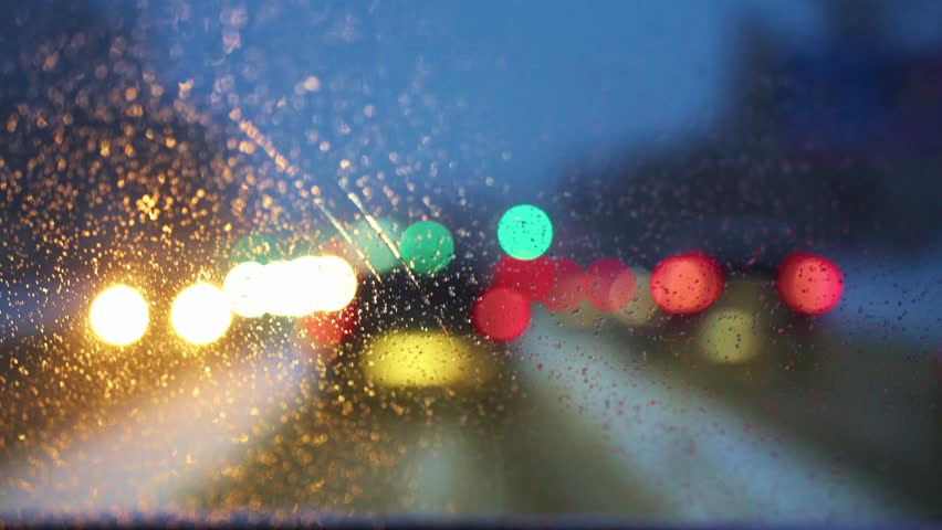 Rain covers the windshield, creating a blurred effect as the driver navigates a busy city street. Bright lights from surrounding vehicles and traffic signals blend together in a vibrant display of