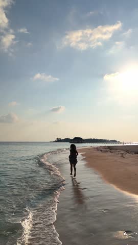 woman runs along the tropical beach at sunset with long shadows cast on the sand, surrounded by calm waves and soft golden light, summer vacation, traveling, family resort, happiness, 