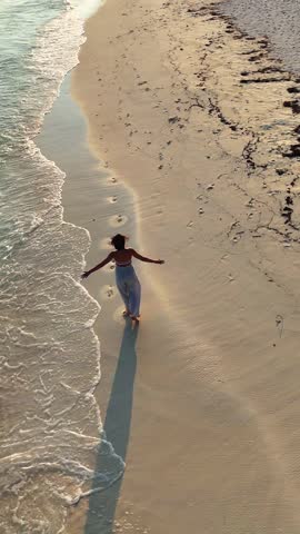 woman runs walks along the tropical beach at sunset with long shadows cast on the sand, surrounded by calm waves and soft golden light, summer vacation, traveling, family resort, happiness, 