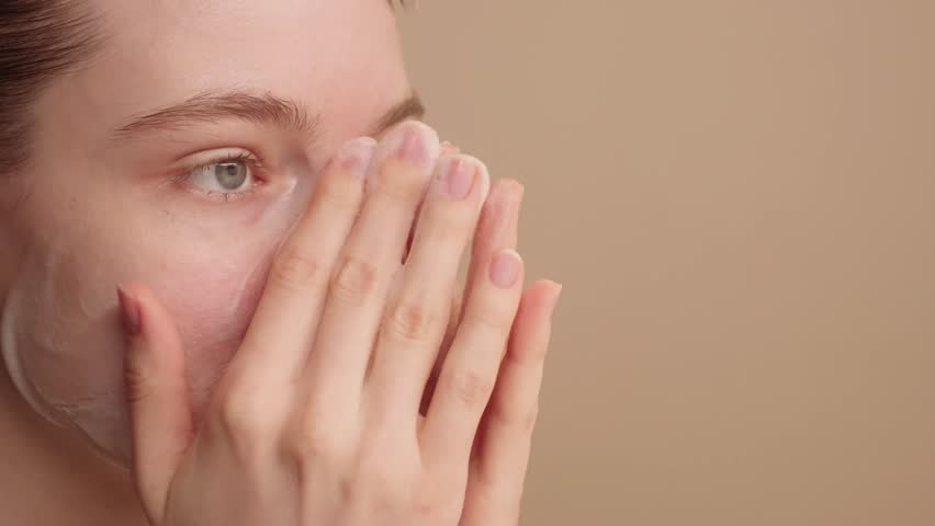 Young beautiful woman gently applying cleansing foam, washing face, daily skincare routine for healthy radiant skin. Advertising concept for daily beauty care on a light studio background. Copy space.