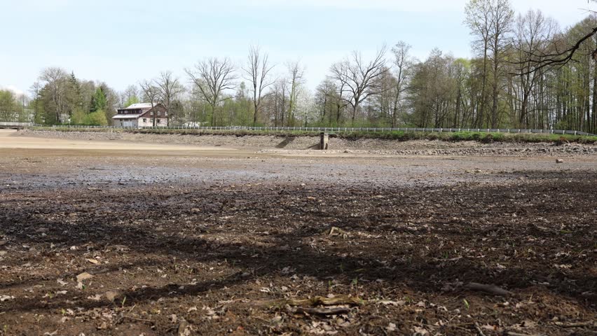 Dry lakebed stretches wide under clear blue sky cracked muddy ground and scattered rocks reveal dramatic drought landscape with forest and distant house in the background