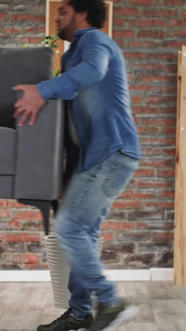 African-American friends carry stylish comfortable sofa in apartment. Men sit down on new couch in living room. Buying furniture for renovated house