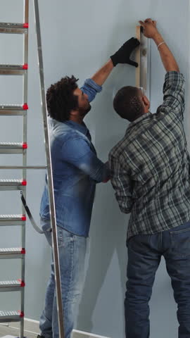 African-American constructors check wall with spirit level working in studio backside view. Black men builders in casual clothes repair home. House renovation