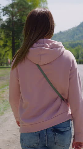 Long haired woman in pink hoodie walks along rural road at eco resort backside view. Tourist spends vacation at highland. Wild nature conservation