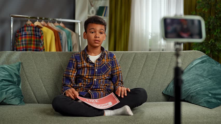 Daily lifestyle concept. Young african american child boy spending time at home, kid blogger records video on smartphone, talking at camera shows paper sheet with like and subscribe text. Slow motion.