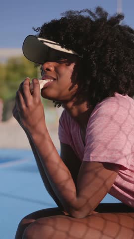 Afro american sportswoman sitting courtside, enjoying quick protein bar, sunlight casting dramatic shadows from fence across pink sportswear during peaceful afternoon break