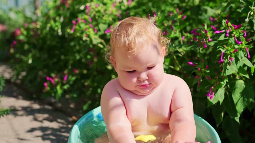 Baby plays with colorful toys in a blue tub surrounded by vibrant flowers on a sunny day in the garden