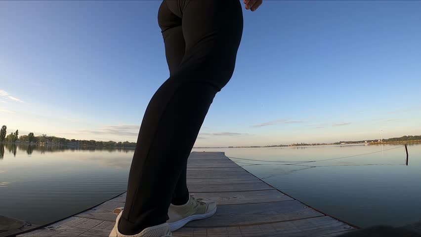 A woman in a black jumpsuit walks along a wooden platform and sits down on it The wooden platform is on the shore of a lake, and there is a beautiful spring sunrise above the lake