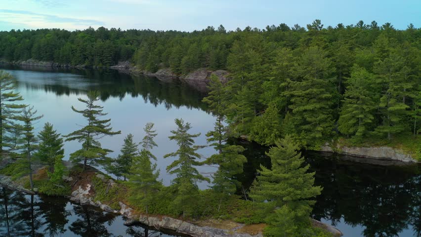 Aerial tracking shot passing a thin island on a calm lake, reflecting the trees on the water during sunset.