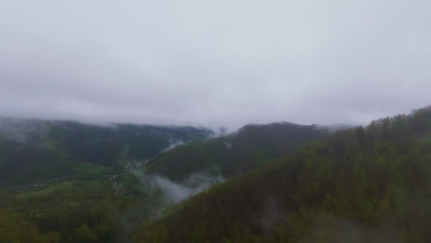 Aerial view of misty green mountains covered with lush forest, fog drifting through hills under a cloudy sky, creating a beautiful natural landscape perfect for travel, adventure, and nature tourism