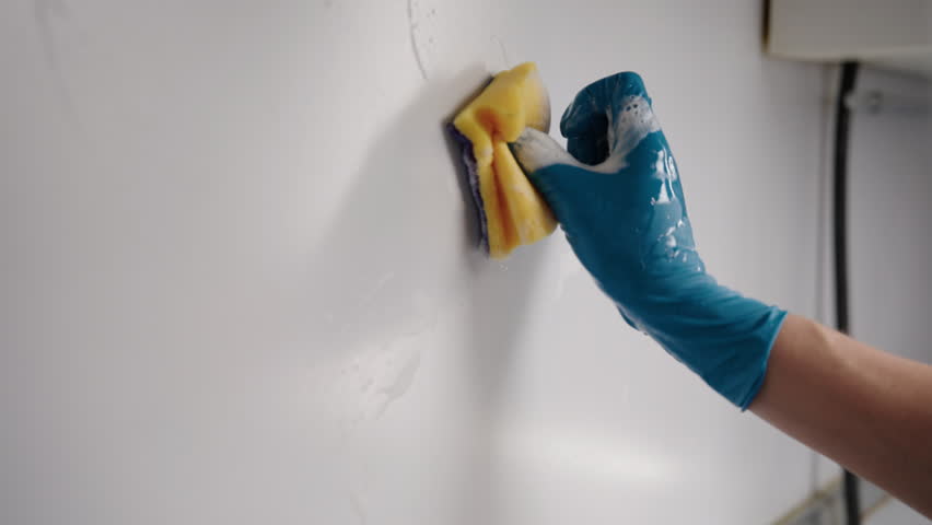 Professional cleaning service worker in blue glove scrubbing white kitchen wall with yellow sponge, creating soap foam while ensuring cleanliness and hygiene in home environment, slow motion, close-up