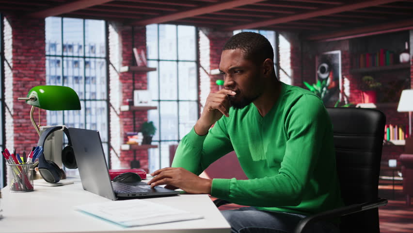 Black man doing remote work feeling uneasy and anxious at desk, shaking his legs and being jittery due to task completion and deadline. Freelancer working under pressure, anxiety. Camera B.