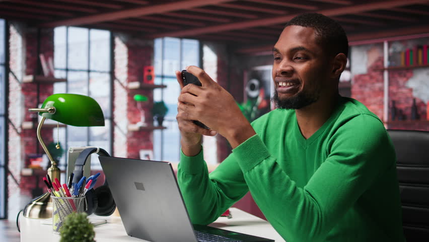 Man at home using smartphone to compose social media posts, producing content as a freelancer. African american guy monitoring profile engagement using mobile phone. Camera B.