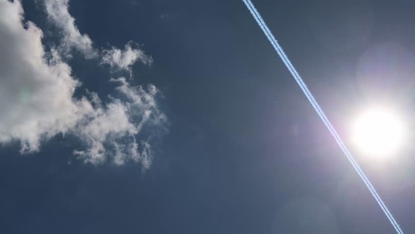 Bright sunlight creating dramatic lens flare near airplane contrail stretching across pristine blue sky with scattered white clouds