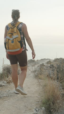 Vertical video. A Woman Balances Along a Narrow Cliffside Path Overlooking the Ocean. Her Arms Are Outstretched for Stability as She Walks Close to the Edge.