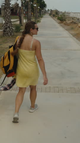 Vertical video. A Woman Walks Away Down a Paved Path, Wrapped in a Yellow Towel, With a Backpack and Shirt in Hand After a Swim.