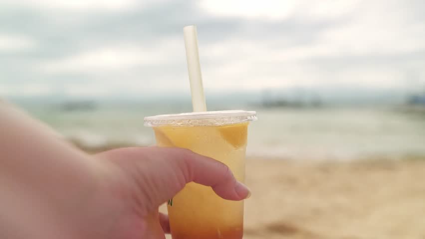 Close-up of hands inserting straw into sealed cup of iced bubble tea at beach with ocean in background, capturing refreshing summer beverage moment in tropical vacation setting. iced bubble tea