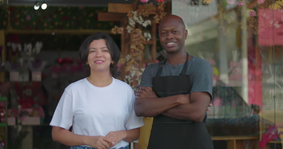 Small business owners standing confidently outside flower shop, smiling man with arms crossed beside woman in white shirt, storefront in background