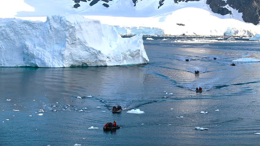 A group navigates through the calm, icy waters of Antarctica in a small boat, marveling at the majestic icebergs under a clear sky. The stunning landscape offers a breathtaking backdrop.