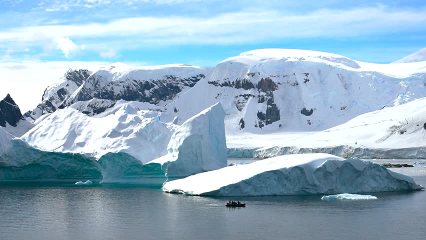 A group navigates through the calm, icy waters of Antarctica in a small boat, marveling at the majestic icebergs under a clear sky. The stunning landscape offers a breathtaking backdrop.