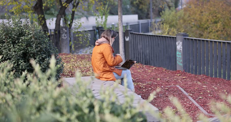 Woman sits on stone surface engaged in online video call via laptop and gesturing with hand. Lady works in autumn cottage garden surrounded with fence