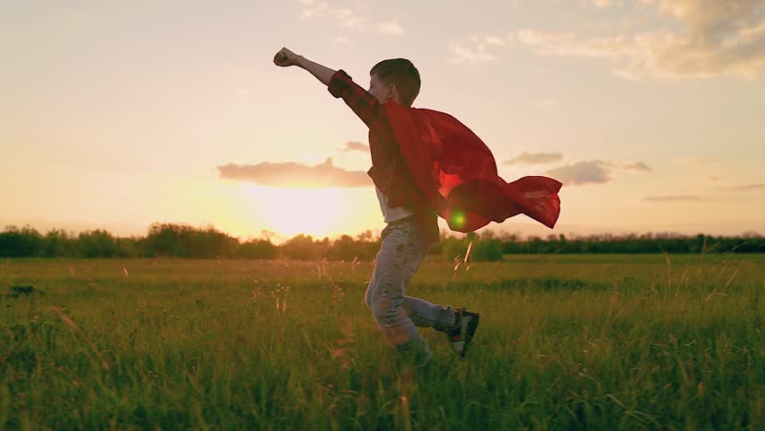 Little boy in red mantle play superhero against sun sky. Kid boy game concept. Child dream, winner runs in superhero costume in nature park. Superhero child boy runs through green meadow in red cloak.