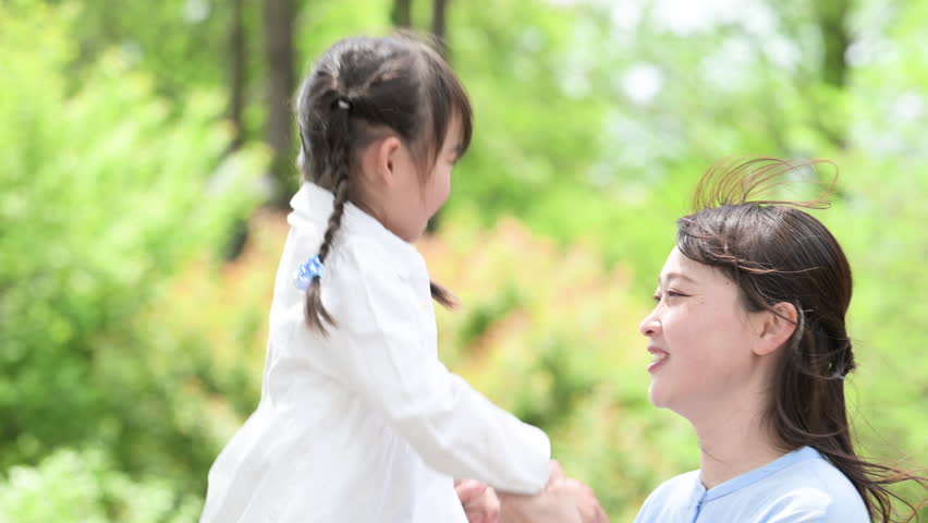 Japanese parent and child (mother and daughter) looking at each other against a background of beautiful fresh greenery. Image of parenting and affection. Close-up video.
