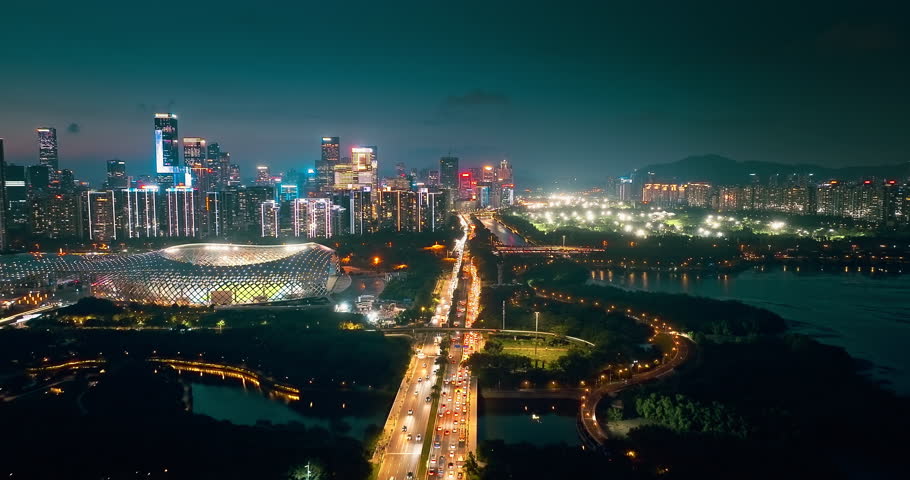 Aerial shot of a modern city district with illuminated stadium, busy highway traffic, and skyline at night in Shenzhen, China.