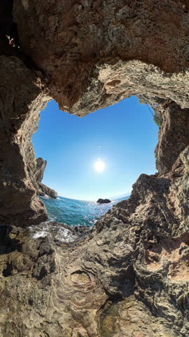 Vertical video. 360 degree camera. A heart-shaped rock formation on the coastline frames a view of the ocean and the bright sun above. Wide-angle shot.