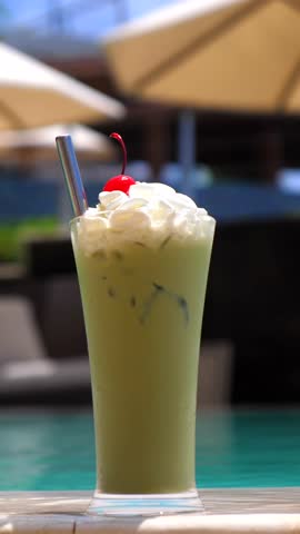 Vertical screen creamy matcha latte topped with whipped cream and a cherry, served in a tall glass with a metal straw, sits poolside under the shade of sun umbrellas