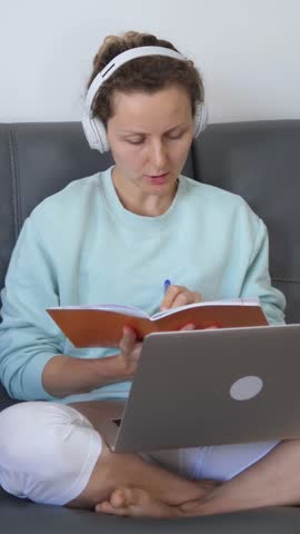Vertical screen comfortable woman wearing headphones sits on a gray sofa, using laptop and taking notes in notebook while participating in online education or remote work