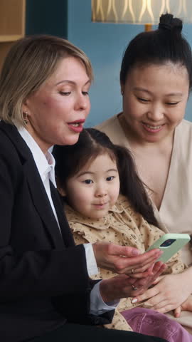 Vertical shot of small Asian girl sitting next to young mom watching mobile phone during visit of Caucasian social worker at home
