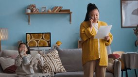 Medium long shot of young Asian mother holding paper talking on mobile phone while little daughter hugging her at home - Powered by Shutterstock - Get 15% off with code: PIKWIZARD15
