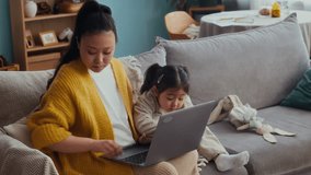 Medium long shot of young Asian mother using smartphone working on wireless laptop while babysitting small daughter at home - Powered by Shutterstock - Get 15% off with code: PIKWIZARD15