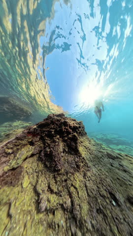 Vertical video. A Man Swims Close to Large Underwater Rocks in Shallow Waters, Light Reflections Creating a Dreamlike 360 View of the Scene.