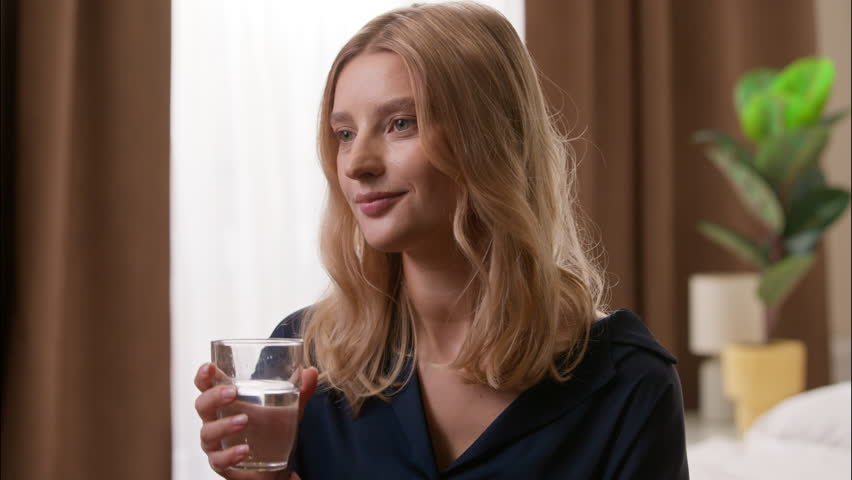 Happy healthy Caucasian lady drinking cup of mineral water refreshing morning in bedroom adult female good habit diet wellness daily routine girl woman holding glass dehydration nutrition healthcare