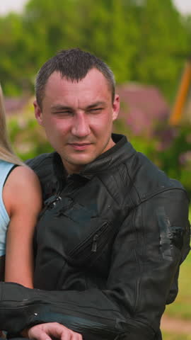 Attractive woman and skilled biker man in leather moto jacket sit on motorcycle against village buildings and gardens on sunny day closeup slow motion