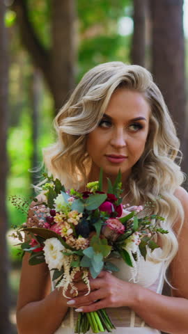 Attractive languid bride with glamorous hairstyle in white transparent wedding dress holds fresh flowers bouquet in city park slow motion
