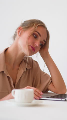 Bored slim blonde woman sits at white table with cup of hot drink in light home office room checking information via modern laptop slow motion
